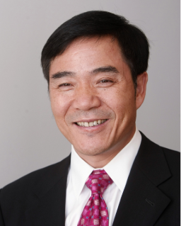 Smiling man with short black hair wearing a black suit, white shirt, and red patterned tie against a neutral background.