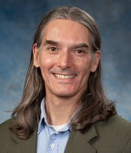 A man with long brown hair, wearing a green blazer and blue collared shirt, smiles at the camera in front of a blue-gray background.