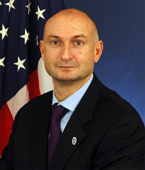 A man in a suit and tie poses in front of an American flag and a blue background.