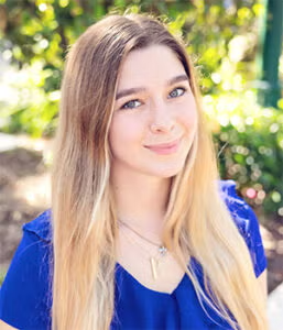 A young woman with long blonde hair wearing a blue top is standing outdoors, smiling at the camera with greenery in the background.