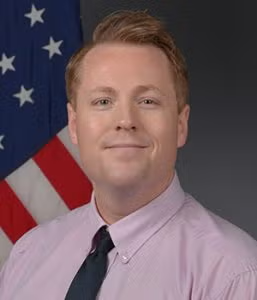 A man in a light purple shirt and dark tie poses in front of a U.S. flag with a neutral background.