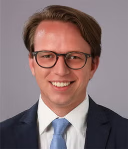 A man wearing glasses, a dark suit, white shirt, and light blue tie, smiling in front of a plain, light gray background.