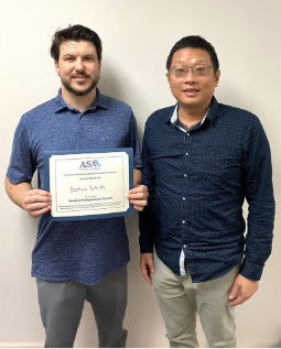 Two men stand side by side; the man on the left holds a certificate with "ASA" at the top, while both face the camera against a plain background.