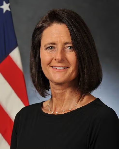 A woman with shoulder-length brown hair wearing a black top stands in front of a U.S. flag, smiling at the camera.