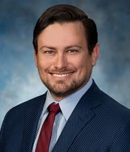 A man with brown hair and a beard wearing a blue suit jacket, light blue shirt, and red tie, smiles at the camera against a blue-grey background.