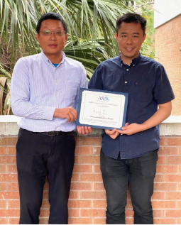 Two men stand in front of a brick wall and greenery; one is holding a framed certificate while the other stands beside him.