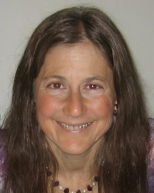A woman with long brown hair, wearing a necklace and earrings, smiles at the camera against a plain background.