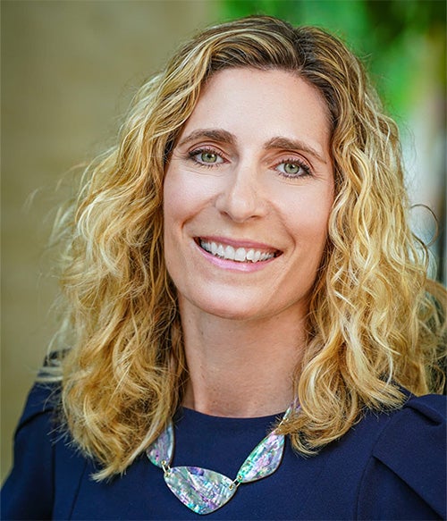 A woman with shoulder-length curly blonde hair, wearing a dark top and a statement necklace, smiles at the camera against a blurred background.