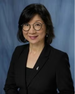 A woman with short black hair and glasses is wearing a black blazer and smiling in front of a blue background.