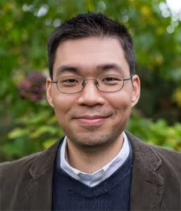 A man with short dark hair and glasses smiles at the camera, wearing a dark jacket and sweater over a collared shirt, with greenery in the background.