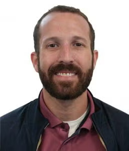 A man with short brown hair and a beard smiles at the camera, wearing a maroon collared shirt and a dark jacket against a white background.