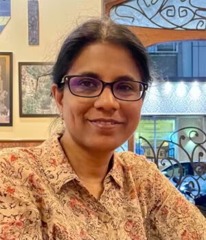 A woman with glasses and dark hair sits indoors, smiling, wearing a patterned shirt with framed pictures and decorative ironwork in the background.