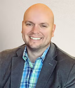 A man with a bald head, smiling, wearing a grey blazer over a blue and green plaid shirt, seated against a light background.