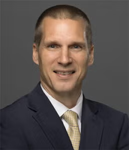 A man in a dark suit, white shirt, and yellow tie smiles at the camera against a plain gray background.