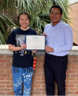 Two people stand outside in front of plants; one holds a certificate while both face the camera and smile.