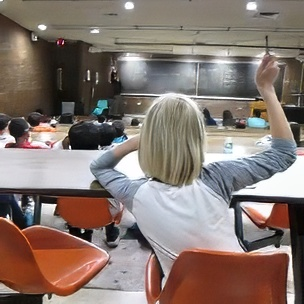 A student with blonde hair raises their hand in a college lecture hall filled with students facing a chalkboard.