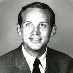Black and white portrait of a man in a suit and tie, smiling, with short hair, facing the camera against a plain background.