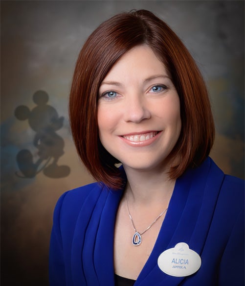 A woman with straight auburn hair wearing a blue blazer and a name tag that reads "Alicia," standing in front of a backdrop with a faint Mickey Mouse silhouette.