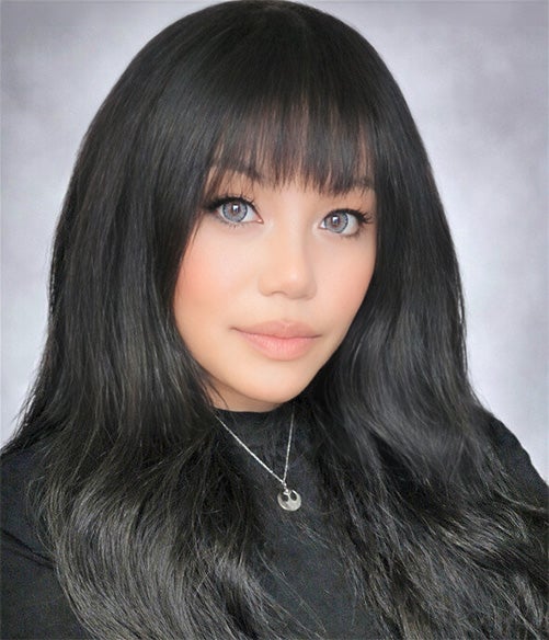 A woman with long, straight black hair and bangs, wearing a black top and a necklace, poses in front of a plain light gray background.