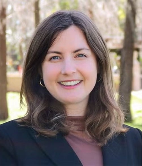 A woman with shoulder-length brown hair, wearing a black blazer and brown top, smiles outdoors with trees and greenery in the background.
