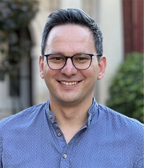 A man with short dark hair and glasses smiles at the camera, wearing a blue patterned shirt, standing outdoors with blurred greenery and buildings in the background.