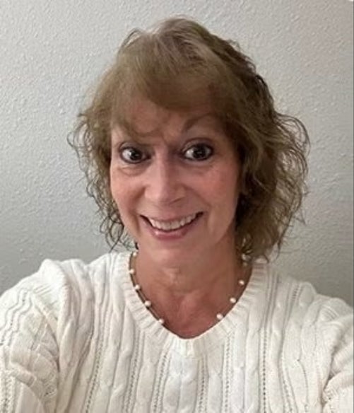 Woman with curly light brown hair wearing a white knit sweater, smiling at the camera, standing in front of a light-colored wall.