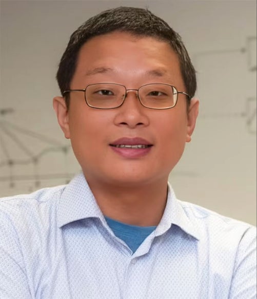 A man with short black hair and glasses, wearing a light blue collared shirt, stands in front of a light background with faint diagrams.