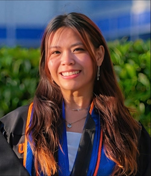 A young woman wearing a graduation gown and stole smiles outdoors, with greenery and a blue building in the background.