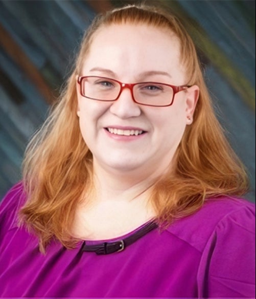 A woman with long reddish-blonde hair, red glasses, and a purple top smiles in front of a blurred, multicolored background.
