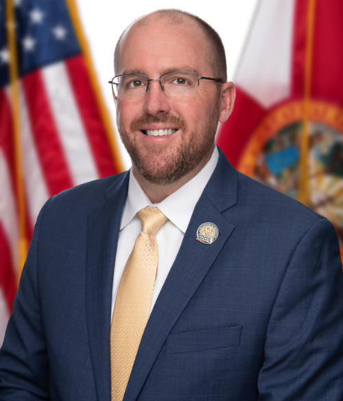 A man in a blue suit and yellow tie stands in front of the U.S. and Florida state flags, smiling at the camera.