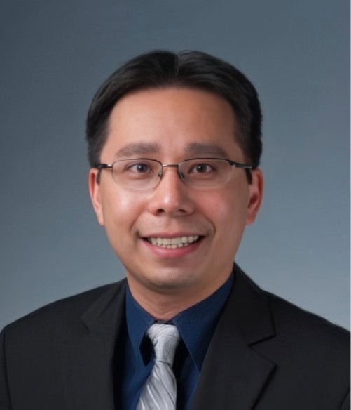 A man with short dark hair and glasses, wearing a dark suit, blue shirt, and patterned gray tie, poses in front of a plain gray background.