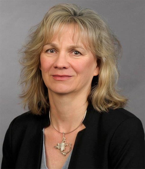 A middle-aged woman with blonde, shoulder-length hair wearing a black blazer, grey top, and a silver necklace, posed against a plain grey background.