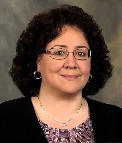A woman with curly dark hair, wearing glasses, hoop earrings, a necklace, and a patterned top with a black cardigan, poses in front of a neutral background.