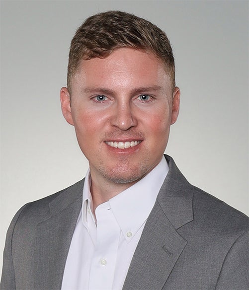 A man wearing a gray suit jacket and white dress shirt, smiling at the camera against a light gray background.
