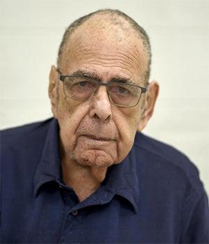 An older man with glasses, short hair, and a serious expression, wearing a dark blue shirt, facing the camera against a plain light background.