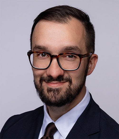 Man with short dark hair, beard, and glasses wearing a dark suit, white shirt, and dark tie, smiling slightly against a plain light background.