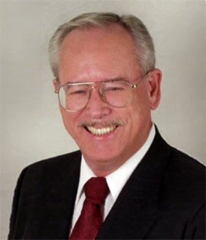 An older man with gray hair, glasses, and a mustache, wearing a dark suit, white shirt, and red tie, smiling at the camera.