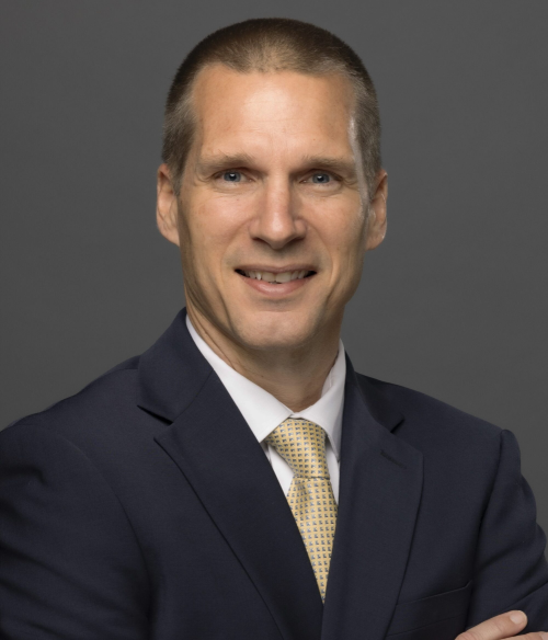 A man in a dark suit, white shirt, and patterned yellow tie poses against a plain gray background with his arms crossed and a slight smile.