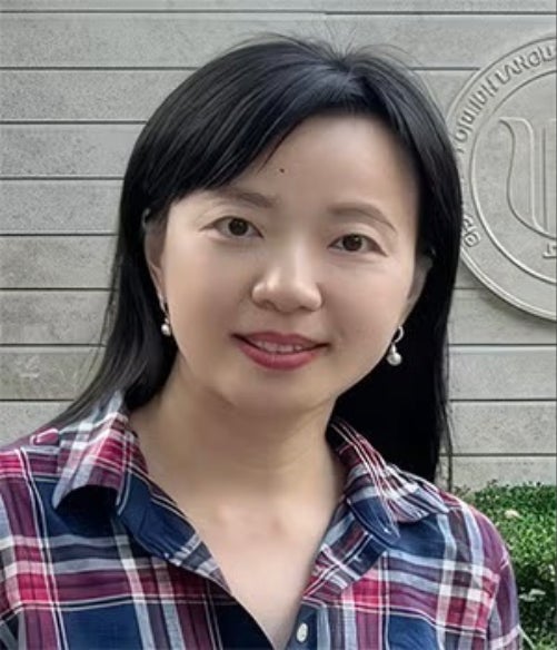 A woman with straight black hair and pearl earrings, wearing a plaid shirt, stands outdoors in front of a stone wall with an embossed emblem.