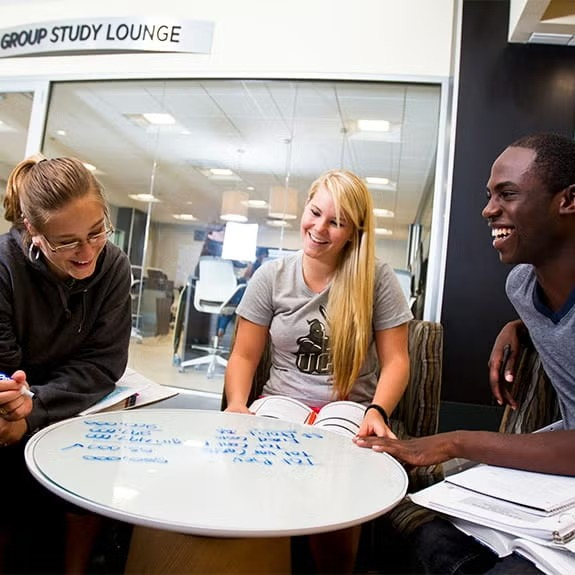 Three students sit around a whiteboard table covered in notes, smiling and studying together in a group study lounge.