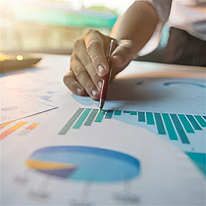 A person points at a bar graph on a document with a pen. Other charts and papers are visible on the table.