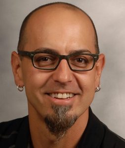 A man with a shaved head wearing glasses and earrings smiles against a gray background. He has a goatee beard.