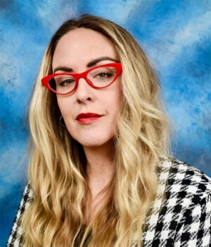 A woman with long wavy blonde hair, red cat-eye glasses, and red lipstick poses against a blue mottled background. She is wearing a black and white checkered jacket.