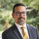 A man with short dark hair, glasses, and a goatee wears a dark suit, white shirt, and yellow striped tie, standing outdoors with greenery in the background.