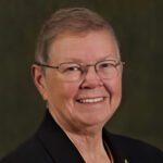 A person with short gray hair, glasses, and a dark suit jacket smiles at the camera against a dark background.