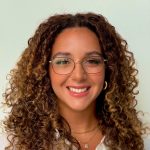 A smiling young woman with curly hair wearing glasses and a white shirt against a plain background.