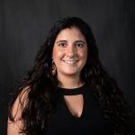 A woman with long, wavy dark hair is smiling at the camera. She is wearing a black sleeveless top with a keyhole neckline and gold earrings. The background is dark and plain.