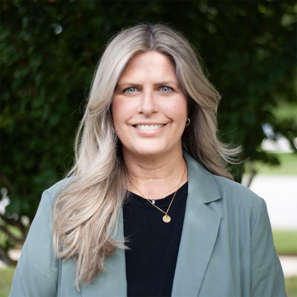 A woman with long, light blonde hair wearing a green blazer and black top stands outdoors in front of leafy greenery, smiling at the camera.