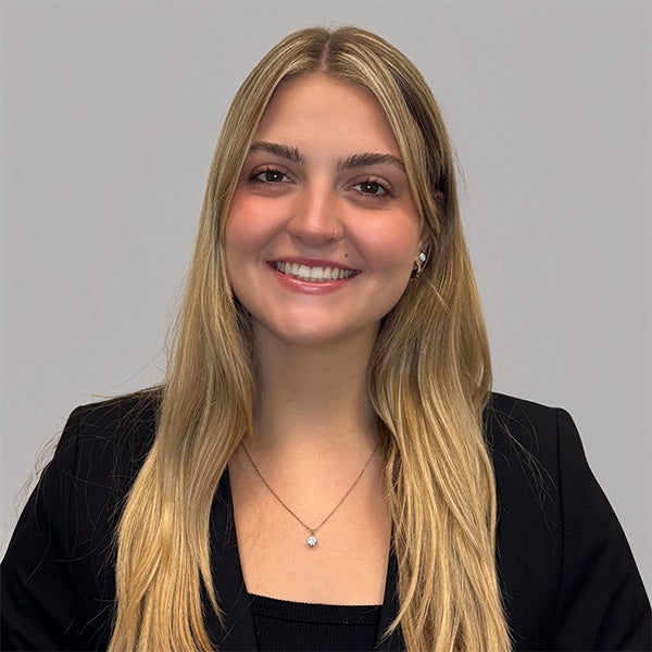 A woman with long blonde hair, wearing a black blazer and necklace, smiles at the camera against a plain light gray background.