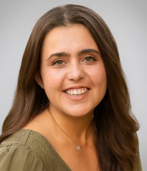 A woman with long brown hair, wearing a green blouse and a necklace, smiles at the camera against a plain light gray background.